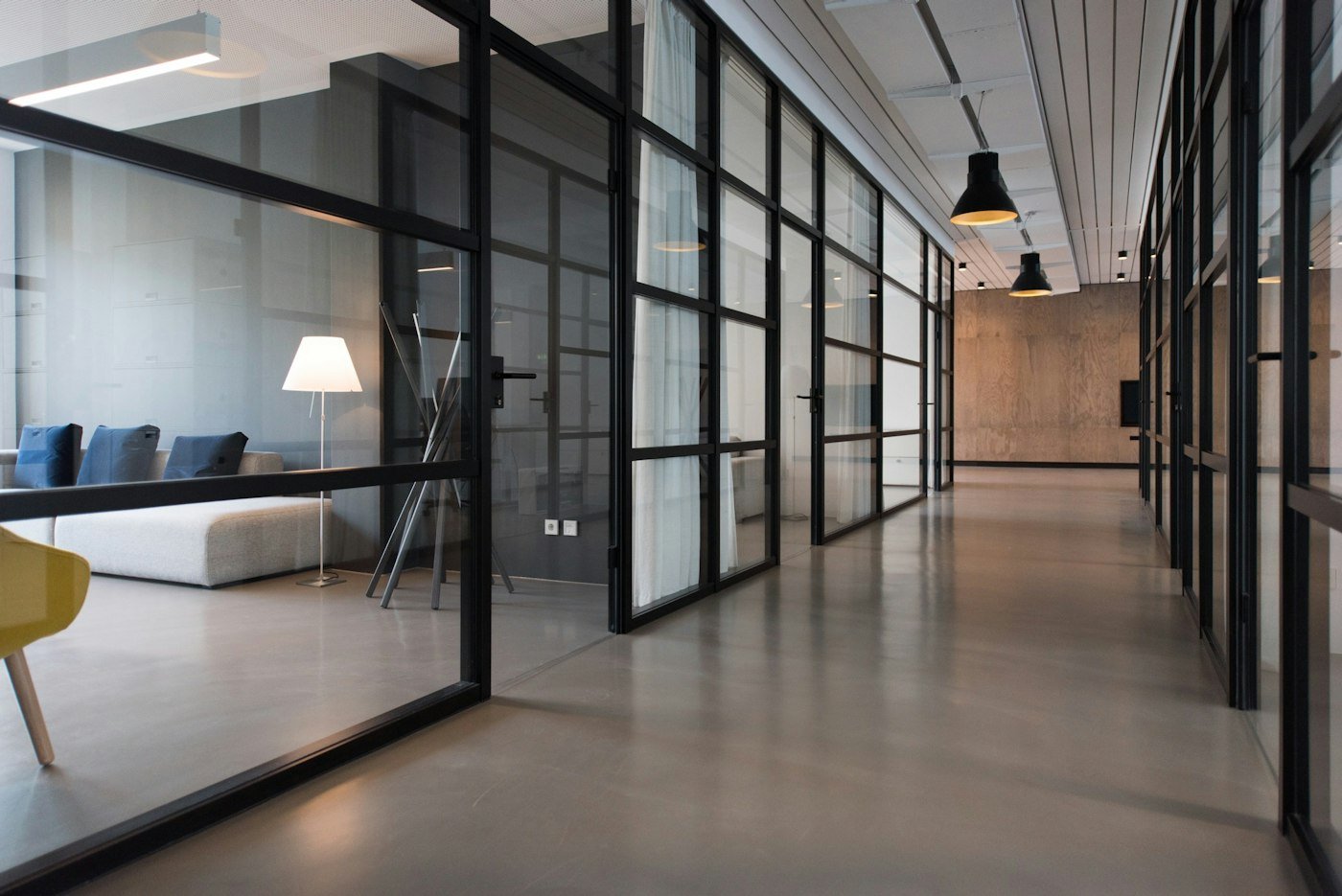 A long shared corridor with glass walls, black frames, a pale floor, and pendant lights